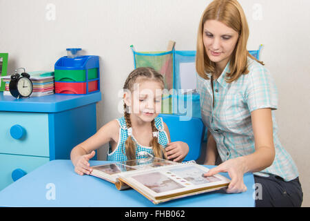 Adulto giovane bella ragazza mostra cinque anni di ragazza in album di foto seduti al tavolo per bambini a casa Foto Stock