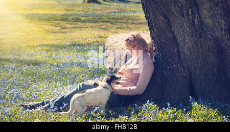 Giovane donna leggendo un libro con il cane sotto agli alberi in primavera Foto Stock