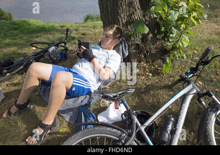 Giovane età bicycler 12 appoggiati su erba con albero come un cuscino controllando il suo telefono cellulare per i messaggi. Rzeczyca Polonia centrale Foto Stock