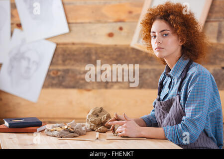 Piuttosto giovane donna potter con ricci capelli rossi seduto e lavoro in studio di ceramiche Foto Stock