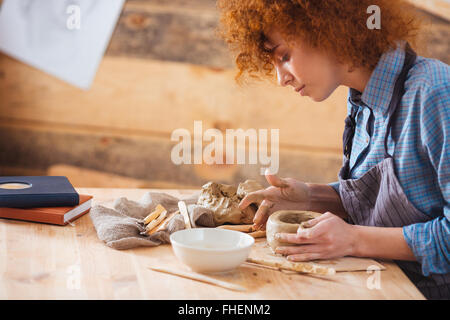 Redhead attraente giovane donna seduta in ceramica studio e creazione di un vaso di terracotta Foto Stock