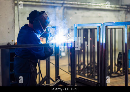 Posteriore lato destro di close-up di un lavoratore che indossa un completo equipaggiamento protettivo, con maschera e guanti di protezione e un grembiule in pelle, saldando insieme Foto Stock