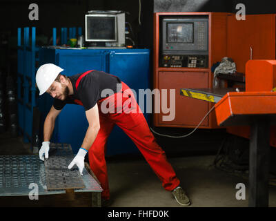 Vista frontale di un lavoratore, indossa una tuta rossa, bianco casco di protezione e guanti, piegamenti e prelevare un foglio di metallo p Foto Stock