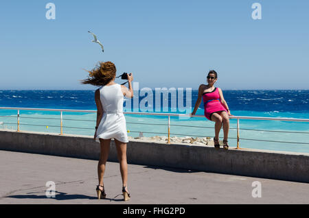 Due amiche scattando foto di ogni altro come essi pongono accanto al mare di Nizza, Francia Foto Stock