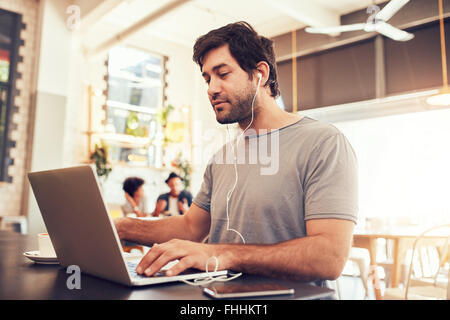 Ritratto di un giovane uomo caucasico con cuffie utilizzando il portatile in un cafe'. Uomo con barba lavorando sul computer portatile a un caffè s Foto Stock