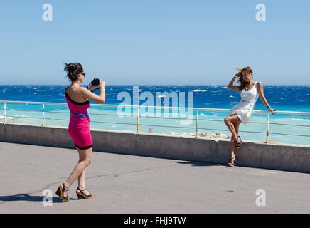 Due amiche scattando foto di ogni altro come essi pongono accanto al mare di Nizza, Francia Foto Stock