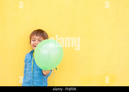 Sorridente ragazzino con palloncino verde e streamer in piedi nella parte anteriore della parete gialla Foto Stock