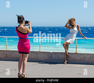 Due amiche scattando foto di ogni altro come essi pongono accanto al mare di Nizza, Francia Foto Stock