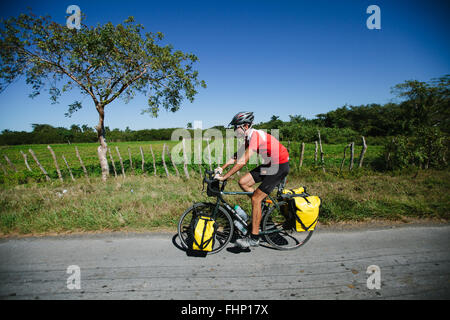 Un uomo in sella ad una bicicletta da turismo caricato con giallo gerle attraverso il paesaggio di Cuba Foto Stock