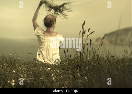 Giovane donna seduta sulla scogliera gesticolando e tenendo l'erba in attesa del suo amante Foto Stock