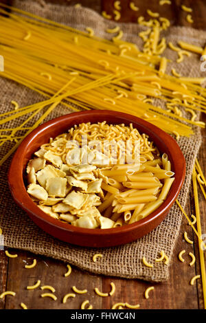 Primo piano di un vaso di terracotta con diverse paste alimentari non cotte come ravioli, spaghetti o mostaccioli, su una tavola in legno rustico Foto Stock