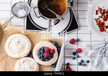 Prodotti da forno freschi per meringa Pavlova dessert di frutti di bosco con lo zucchero in polvere e caramello sull piastrellate di bianco tavolo da cucina. Lay piatto Foto Stock