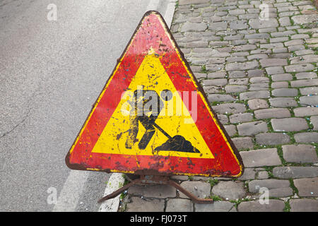 Triangolo rosso e giallo cartello stradale su strada urbana. Gli uomini al lavoro, su strada in costruzione Foto Stock