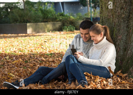 Coppia giovane in autunno park seduta vicino ad albero di ebook di lettura Foto Stock