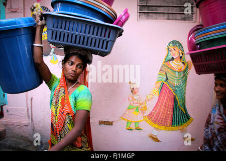 La donna a piedi in una strada di Udaipur, cestello di trasporto sul suo capo, la pittura di una donna con bambino sulla parete, Rajasthan, India Foto Stock