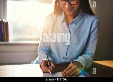 Messa a fuoco selettiva su mani di donna sorridente in blu camicetta iscritto al desk sul blocco note in home office con la luce del sole su di lei dovrebbe Foto Stock