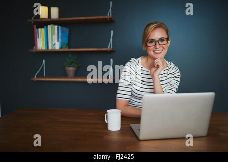 Donna con notebook sorridente in telecamera seduta in ufficio a bere caffè, colori blu Foto Stock