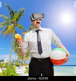 Giovane imprenditore visualizzando una spiaggia ambiente utilizzando una cuffia VR e tenendo un cocktail e una palla spiaggia Foto Stock