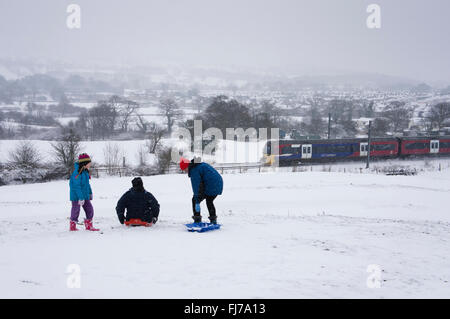 Campagna invernale scena - nella neve, una famiglia di 3 persone con slitte, mentre un treno viaggia al di là del passato - Burley in Wharfedale, West Yorkshire, GB, UK. Foto Stock