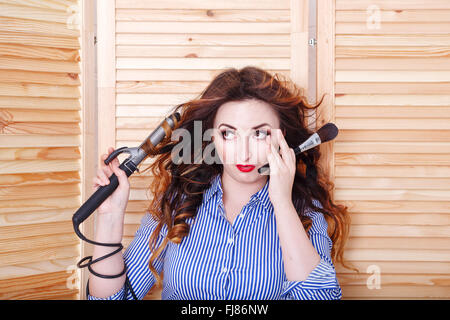 Giovane ragazza attraente con arricciacapelli e pennelli per il trucco. Ragazza in una camicia a righe e tacchi alti. Capelli lunghi. Foto Stock