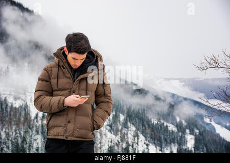 Dai capelli scuri bel giovane in inverno cappotti tramite telefono cellulare o smartphone, outdoor in montagna con paesaggio innevato behi Foto Stock