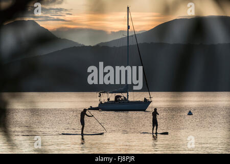 Silhouette di 2 stand up paddle boarders al tramonto lungo le coste di un paradiso tropicale isola in acque calme con barche a vela in background. Foto Stock