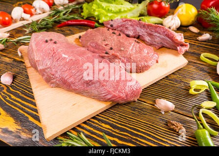 Tre materie a base di carne rossa tagli sulla tavola di legno e tavolo circondato da acqua calda e piccante di verdure e condimenti Foto Stock