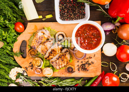 Angolo alto ancora vista la vita di grigliate di carne di maiale o di pollo trita appoggiato sul tagliere di legno con una ciotola di salsa di erbe fresche e Foto Stock