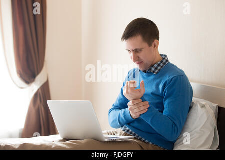 Ritratto di giovane ha sottolineato uomo seduto sul letto con il computer portatile smart di indossare un abbigliamento informale, toccando il suo polso dolente con accorata Foto Stock