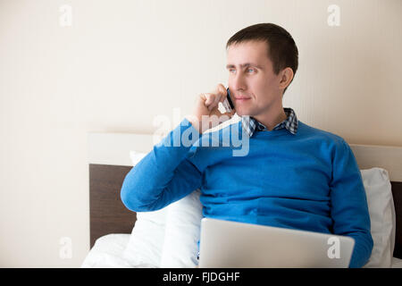 Ritratto di attraente sorridente giovane uomo seduto sul letto con il computer portatile smart di indossare un abbigliamento informale, maschio modello parlando Foto Stock