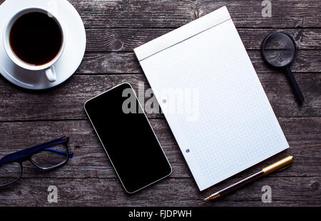 Luogo di lavoro piatta da laici al di sopra di vista. Aperto il blocco note, telefono, occhiali, una lente di ingrandimento su una tavola in legno rustico. Il lavoro moderno env Foto Stock
