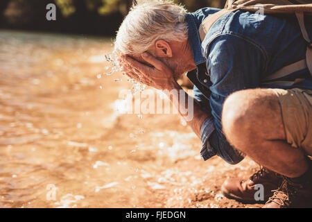 Lato ritratto di un uomo maturo il lavaggio del suo volto nel lago durante un'escursione. L'uomo anziano con zaino seduti a bordo dell'acqua un Foto Stock