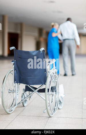 Vista posteriore di infermiere aiutare il paziente a piedi con la sedia a rotelle in primo piano Foto Stock