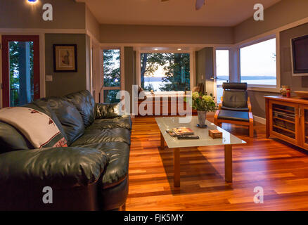 Contemporaneo ed elegante home soggiorno con pavimenti in legno, divano in pelle e un tavolo da caffè, finestra sedile e finestre con vista sull'acqua Foto Stock