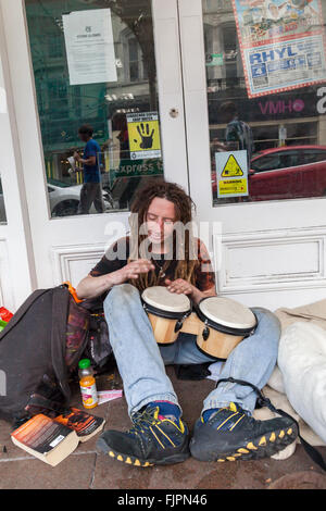 Un giovane maschio caucasico Busker con lunghi dreadlocks seduta sul marciapiede di suonare il tamburo bongo al di fuori di un negozio porta Llandudno Foto Stock