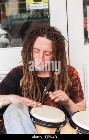 Un giovane maschio caucasico Busker con lunghi dreadlocks seduta sul marciapiede di suonare il tamburo bongo al di fuori di un negozio porta Llandudno Foto Stock