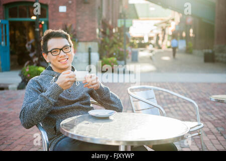 Giovane uomo asiatico seduto e sorridente in un rilassante outdoor cafe azienda tazza di caffè Foto Stock