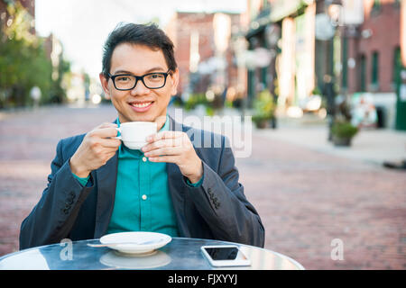 Successo asiatico giovane uomo seduto e sorridente in un rilassante outdoor cafe con telefono mobile holding tazza di caffè godendo il suo bre Foto Stock