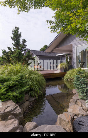 Una funzione di acqua per emulare un torrente scorre sotto un ponte e tra rocce paesaggistici con una casa moderna in background. Foto Stock