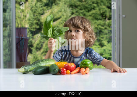 Carino piccolo ragazzo seduto al tavolo, esprimendo emozioni contrastanti su farine vegetali, cattive abitudini alimentari concetto Foto Stock