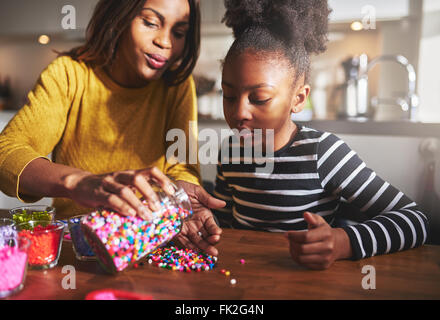 Disponibile madre africana prendendo le perline colorate dal contenitore in mano sul tavolo in legno per bambino seduto in cucina Foto Stock