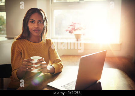 Grave giovane femmina singola seduta a tavola azienda tazza da caffè accanto ad aprire il portatile con il sole luminoso proveniente attraverso la finestra Foto Stock