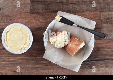 Angolo di alta vista di una cena di sesamo rotoli rotto in metà e imburrato. Accanto alla piastra di pane è un coccio di burro con il coltello. Foto Stock