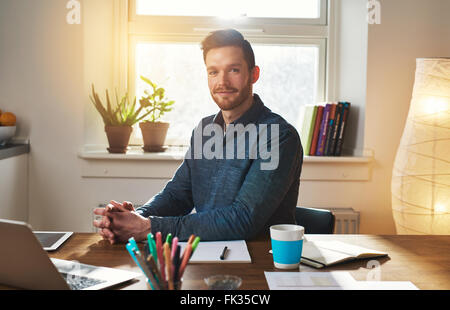 Imprenditore di successo seduto alla sua scrivania nel suo ufficio di casa a pensare come egli guarda la telecamera con un espressione pensieroso Foto Stock