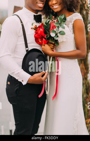 Piuttosto giovane sposa nera e lo sposo felicemente sorridente e tenendo un mazzo di fiori in mezza lunghezza Foto Stock