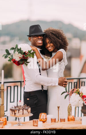 Felice sposa nera ed elegante per lo sposo in posa per la telecamera sulla terrazza con panorama urbano in primo piano Foto Stock