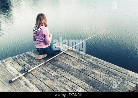 Poco bionda ragazza caucasica seduto su di un molo in legno con canna da pesca, vintage foto stilizzata con correzione delle tonalità effec filtro Foto Stock