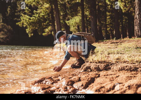 Lato ritratto di senior escursionista maschile seduto da un lago e mettere le mani in acqua. Uomo maturo faccia il lavaggio nel lago. Foto Stock