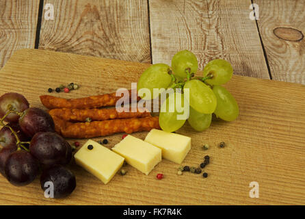 Kabanosy sottile, salsiccia, formaggio ,due grappoli di uva og su un piano di lavoro in legno, ornato di pepe grani colorati. Vista orizzontale da t Foto Stock