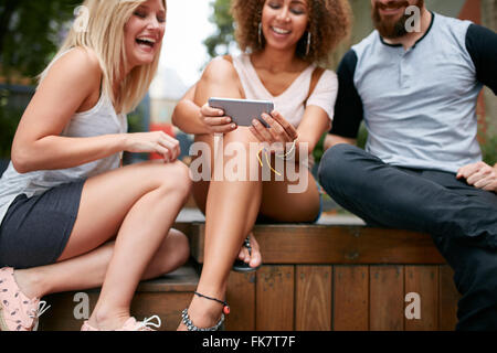 Tre giovani amici guardando la smart phone insieme e ridere. Diversi gruppi di giovani seduti insieme all'esterno lo Foto Stock